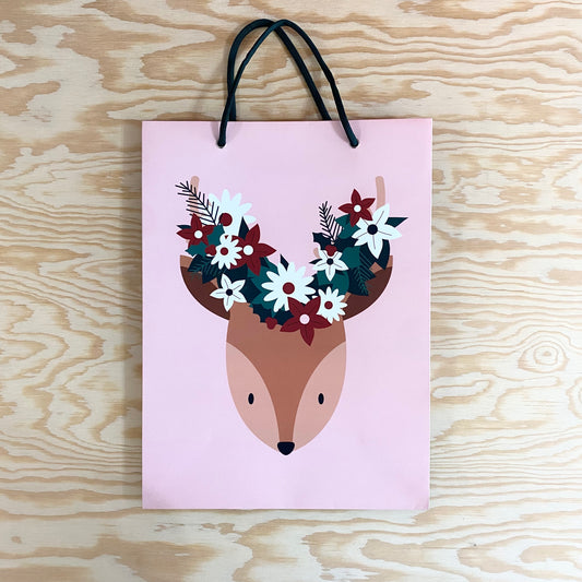 A pink bag with dark green yarn handles. The image of a cartoon deer head with red and white flowers on its head are displayed on the front of the bag.