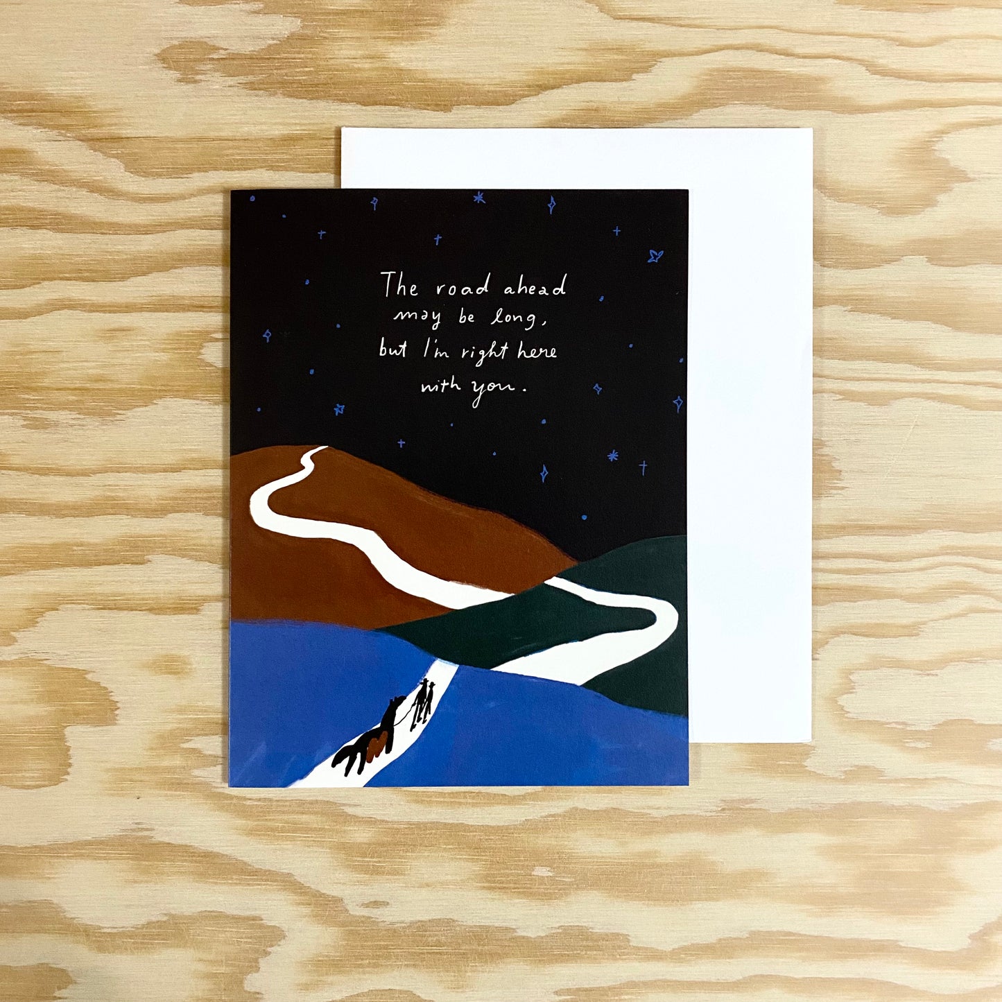 Card with white envelope. Two people with a horse walking on a white path among hills of blue and brown, with a black sky filled with blue stars. The words "The road ahead may be long, but I'm right here with you" are written across the sky.