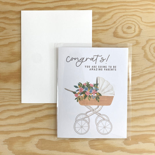 A portrait greeting card with Congrats across the top in cursive. The picture is of a rolling bassinet that has a bouquet of pink and blue flowers in the basket.